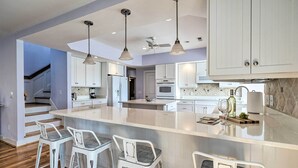 A modern kitchen with white cabinetry, a large island with bar stools, pendant lighting, and a ceiling fan. - Waterfront Hampstead Home: Dock & Waterway Access! (Hampstead)