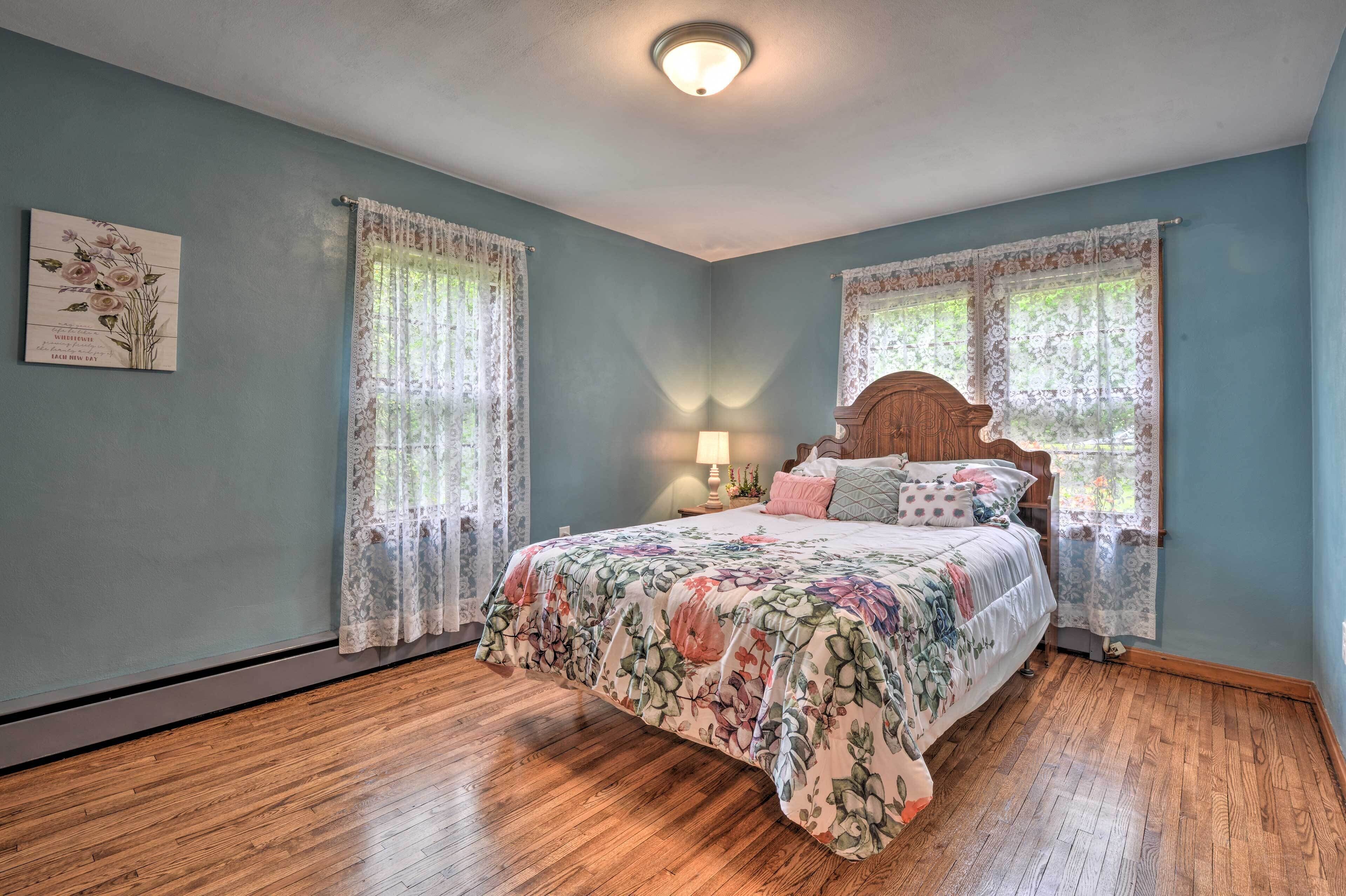A bedroom with a floral bedspread, two windows with lace curtains, a wooden headboard, and a nightstand with a lamp.