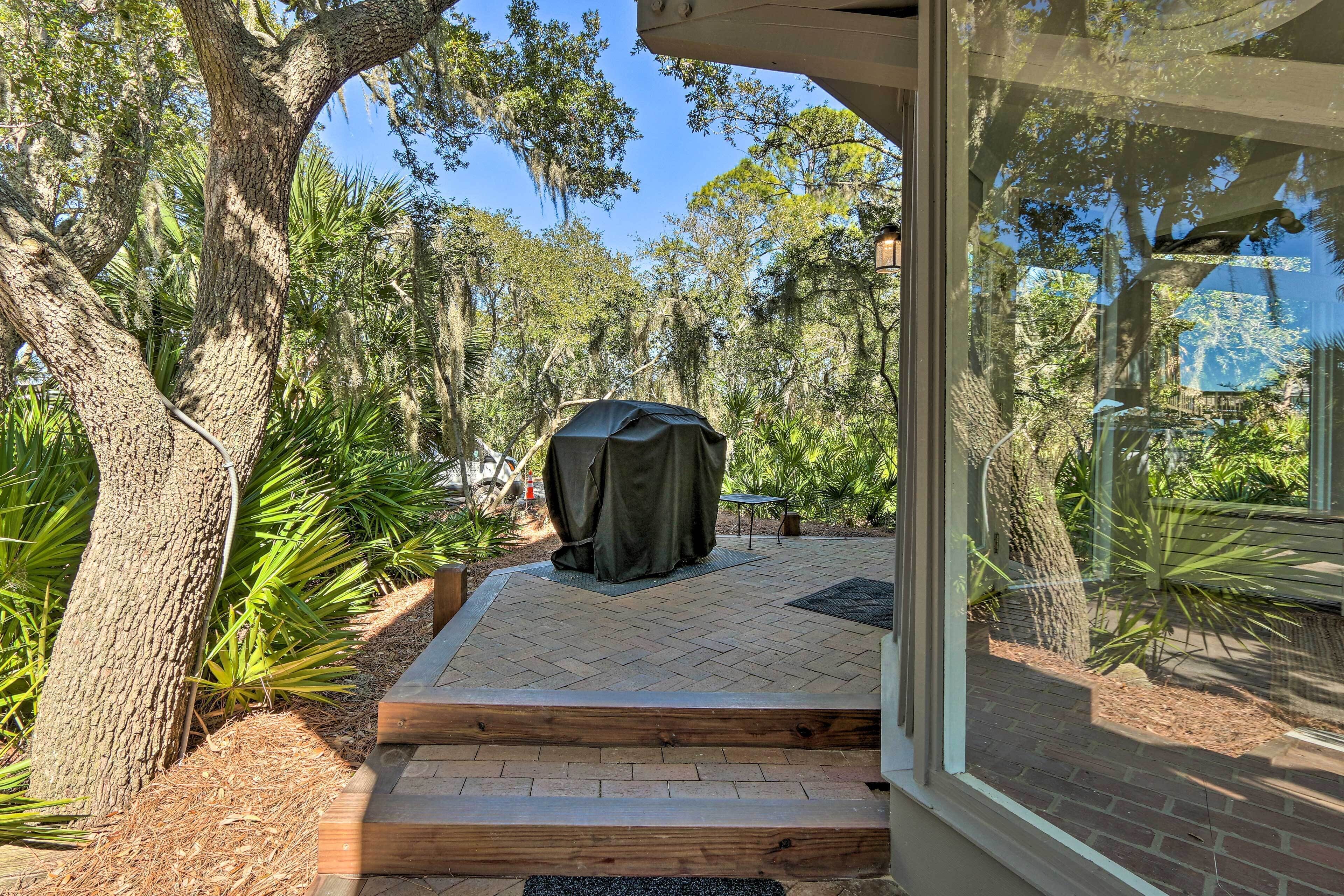 A covered outdoor seating area with steps leading to a glass door, surrounded by trees and plants.