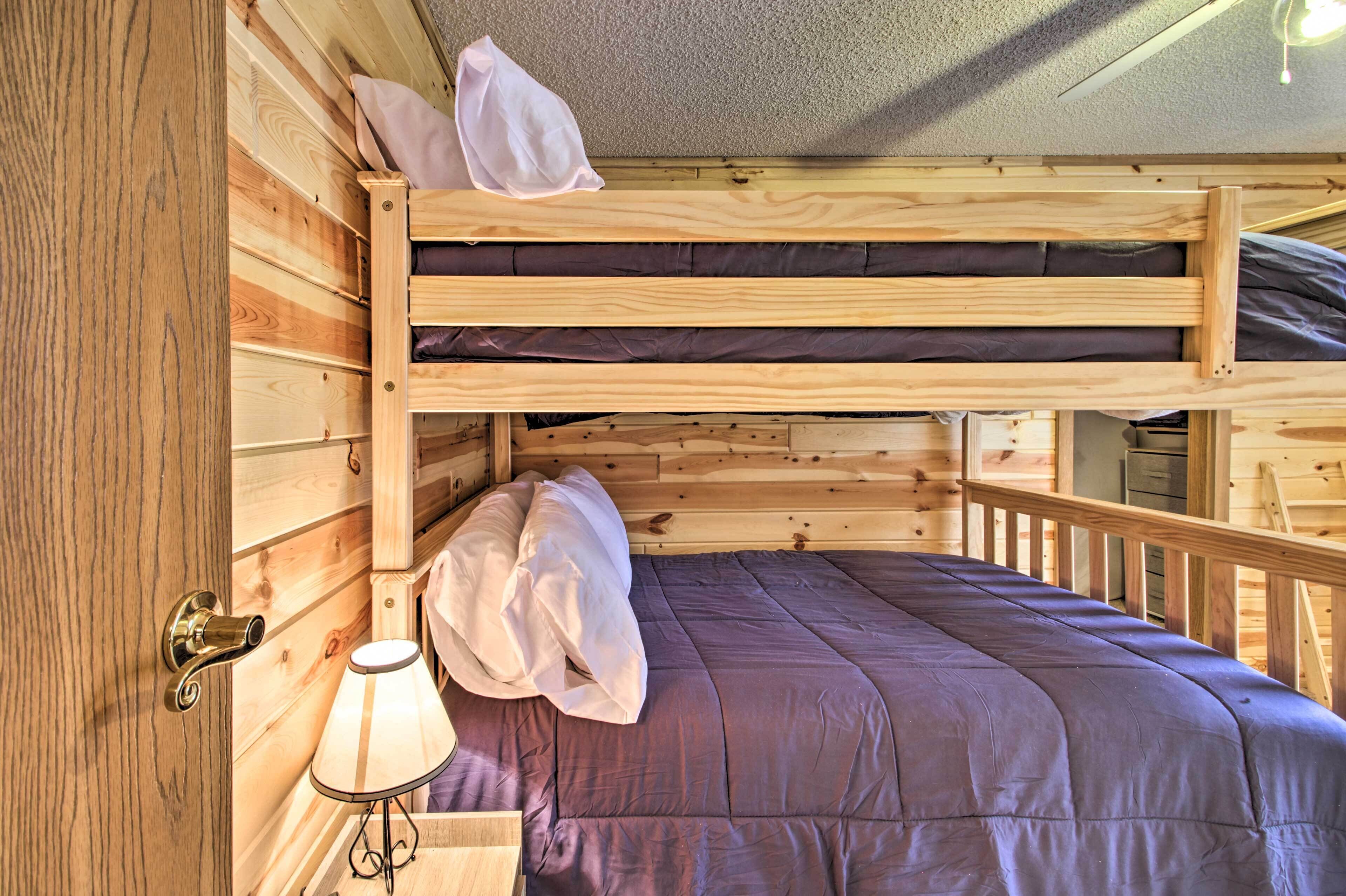 A wooden bunk bed with a purple bedspread and pillows, a small table with a lamp, and a wooden door with a brass handle.