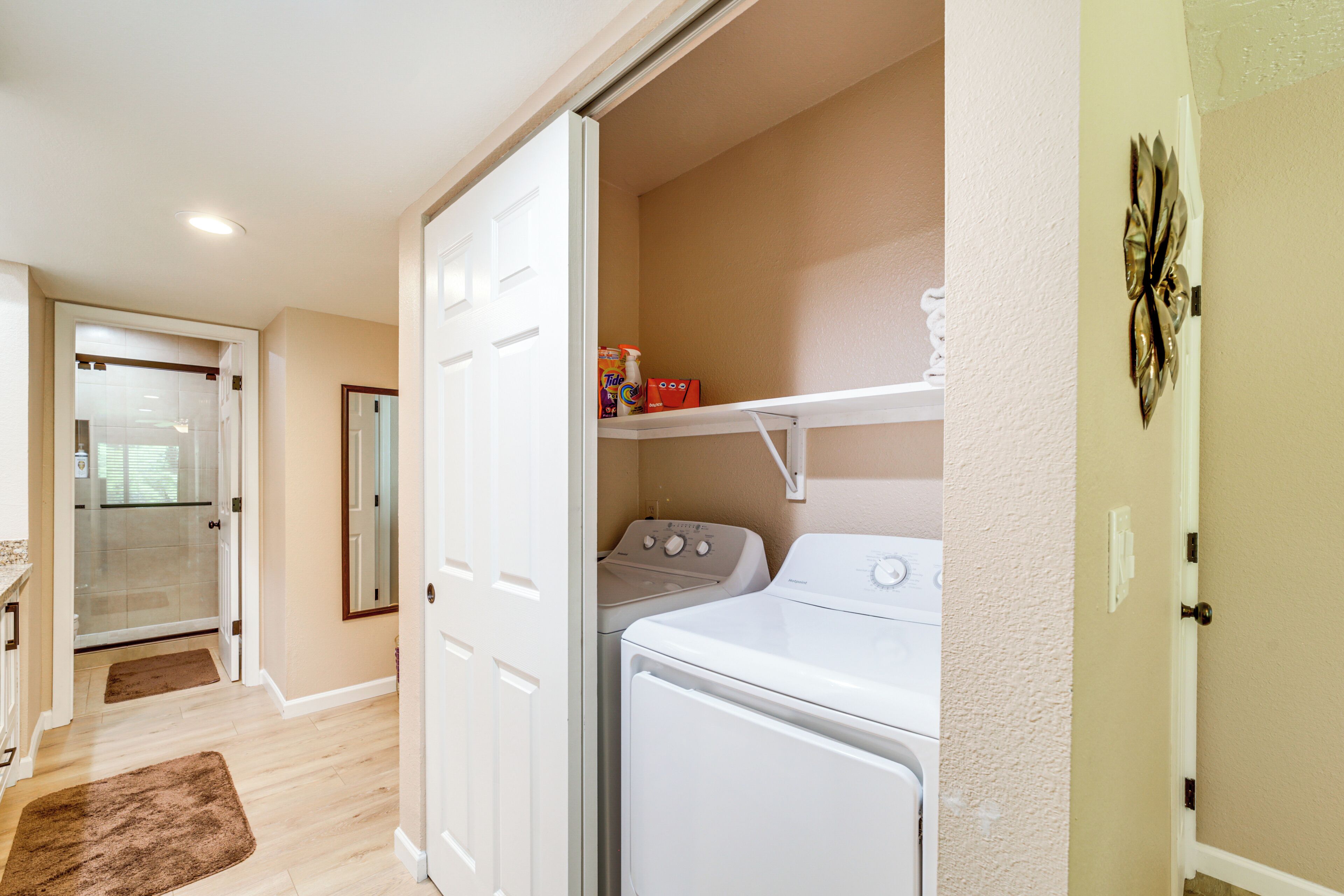 A laundry room with a washer and dryer, a shelf with cleaning supplies, and a door leading to a bathroom.