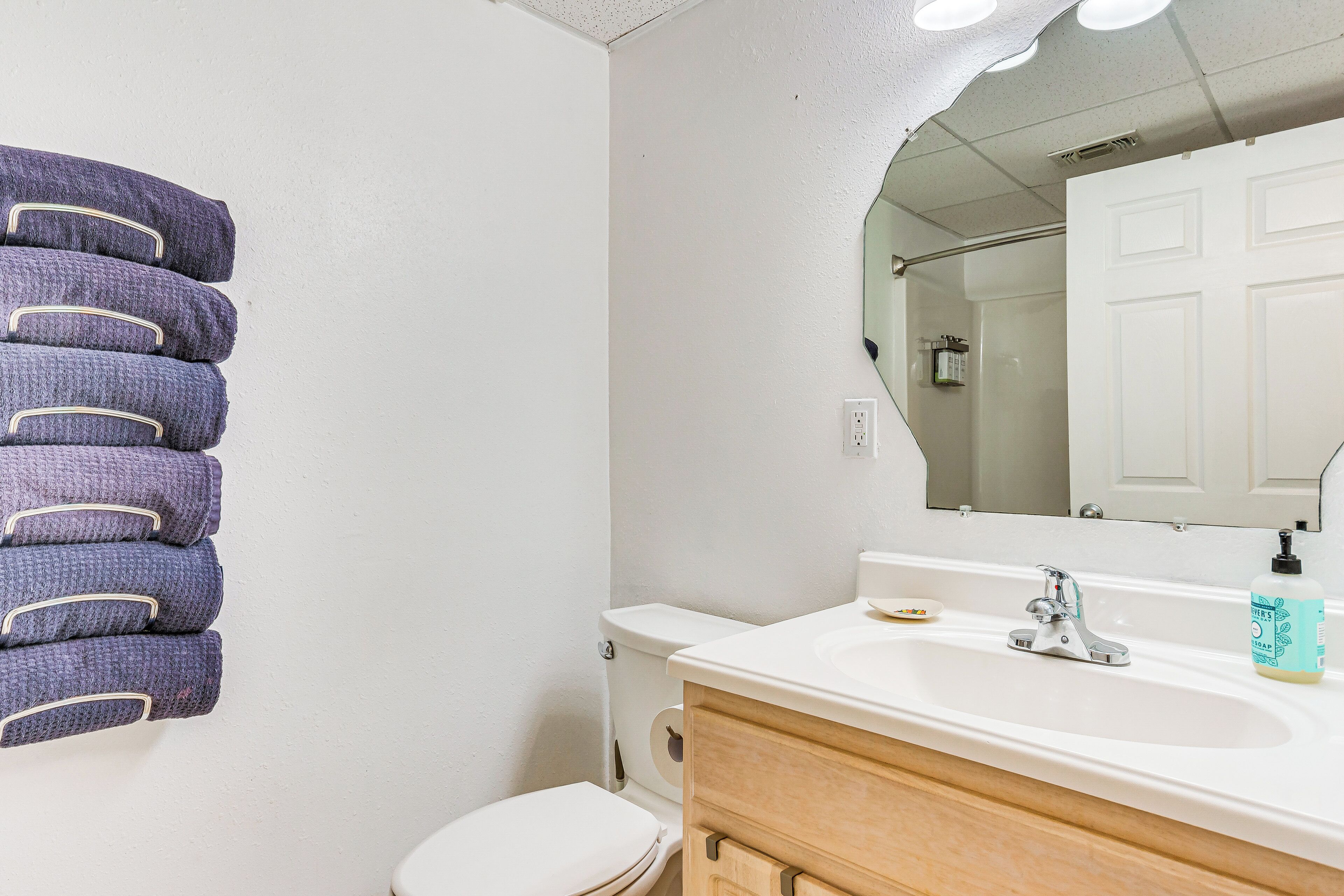 Une salle de bain avec un lavabo blanc, un miroir et des toilettes. Des serviettes sont pliées et empilées sur un porte-serviettes mural.