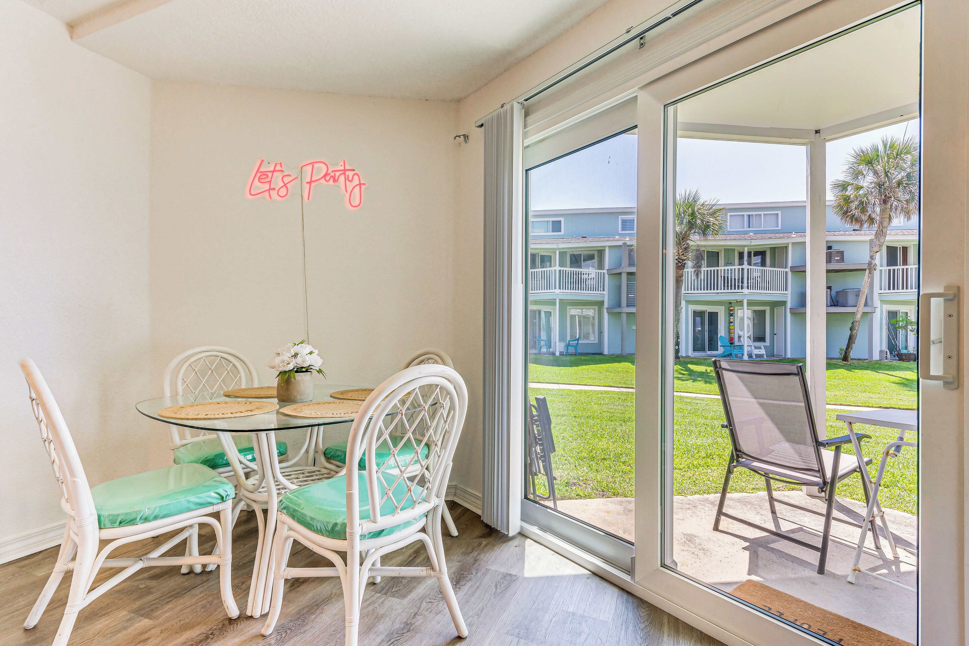 A dining area with a glass table and wicker chairs, a neon sign on the wall, and a view of a residential building through a glass door.