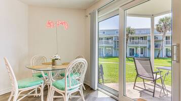 A dining area with a glass table and wicker chairs, a neon sign on the wall, and a view of a residential building through a glass door.