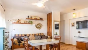 Een woonkamer met een bank, een eettafel met glazen blad, houten stoelen, een plafondventilator, wandplanken en een televisie.