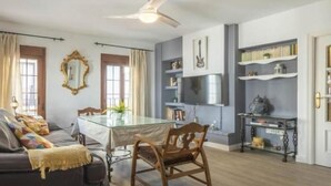 Una sala moderna con una mesa de comedor de vidrio, un sofá, un ventilador de techo y una zona de cocina con estanterías y una estufa.