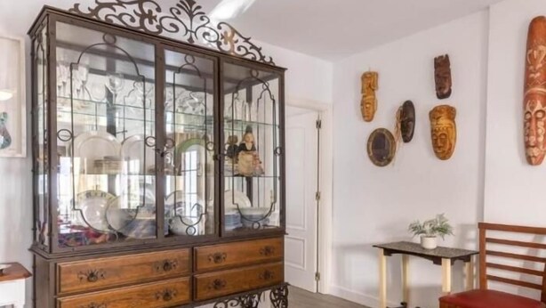 Una habitación con un aparador de madera con puertas de cristal, una mesita con una planta y varias esculturas de madera en la pared.