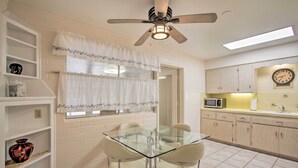 Une cuisine avec une table en verre, des chaises blanches, un ventilateur de plafond, un four à micro-ondes et une horloge au mur.