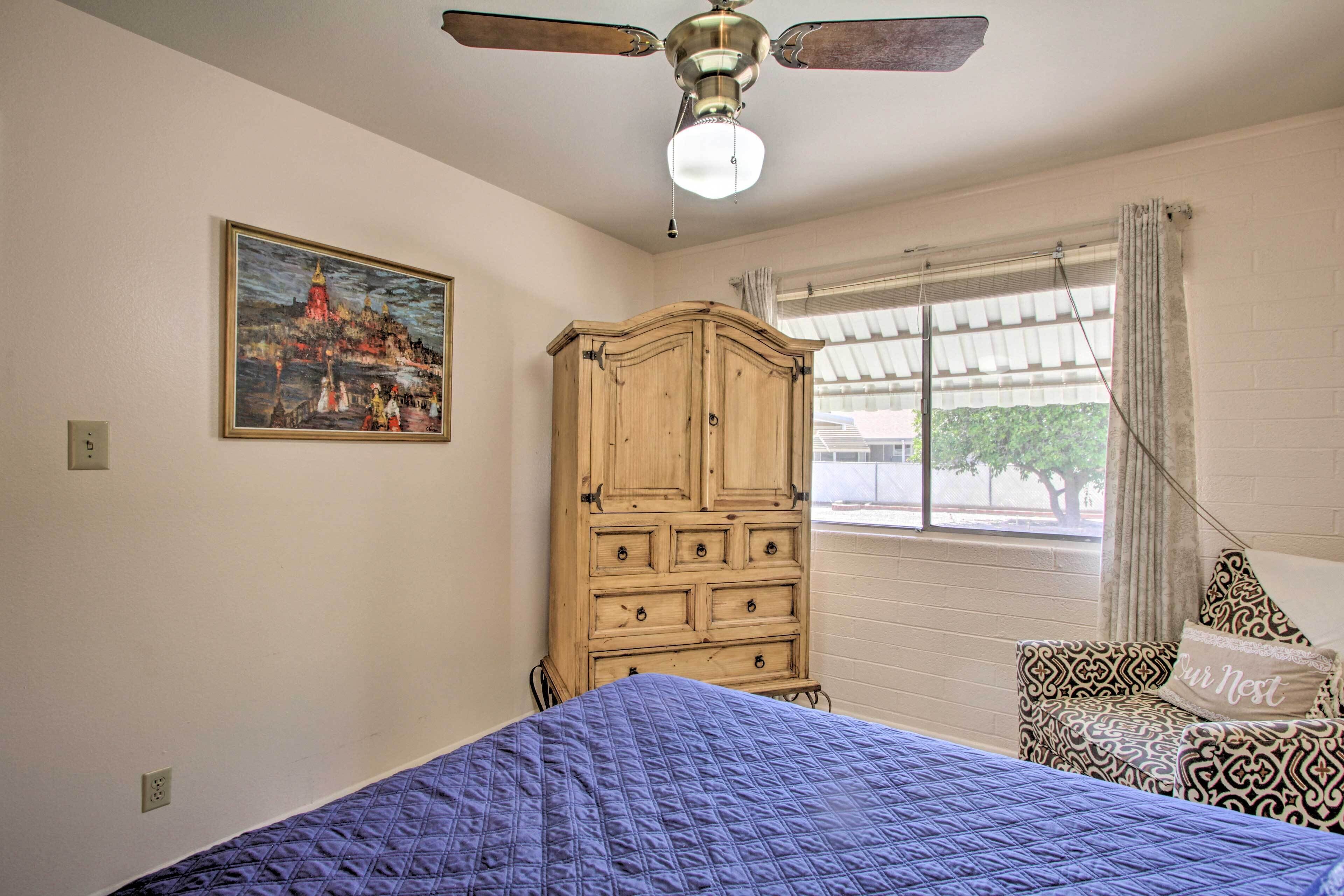 Un dormitorio con cama, ventilador de techo, cómoda de madera, ventana con cortinas y un cuadro en la pared.