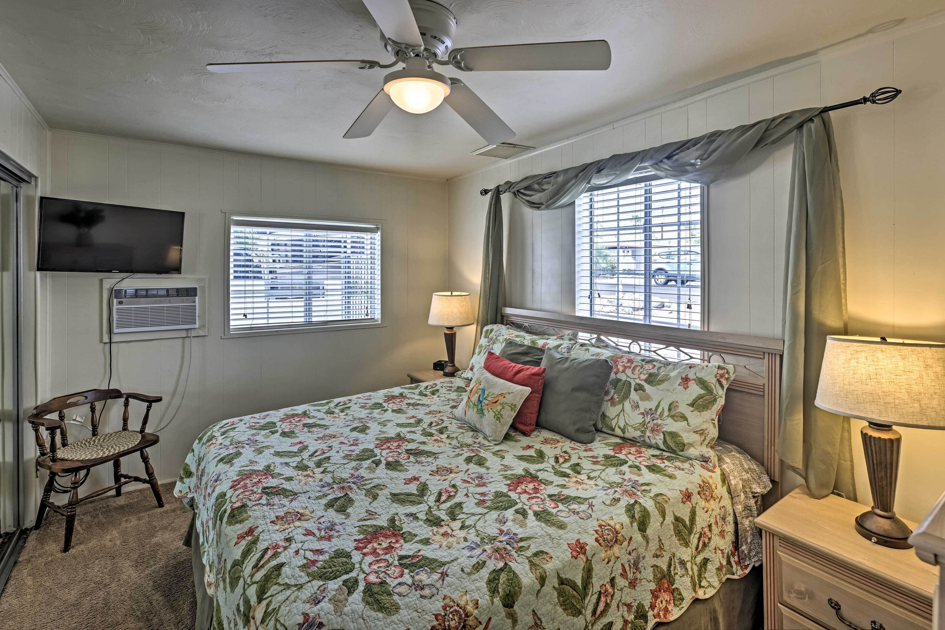 Ein Schlafzimmer mit Bett, Deckenventilator, Fernseher, Fenster mit Jalousien und einem Sessel.