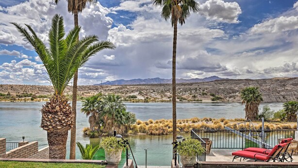 A lakeside patio with palm trees, a red lounge chair, and a view of mountains in the distance. - Restful Riverfront Retreat w/ Private Dock + Patio (Bullhead City)