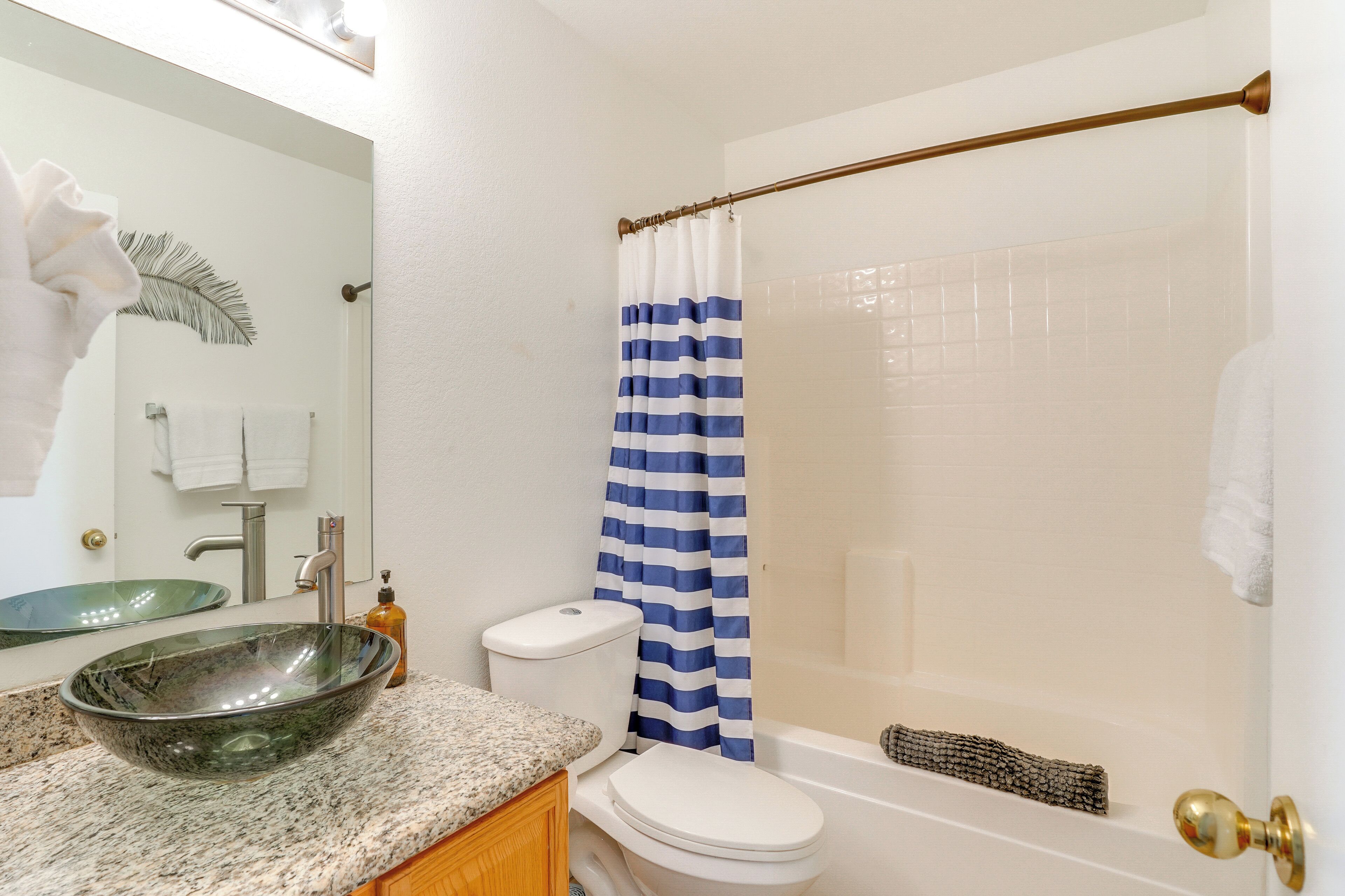 Une salle de bain avec un lavabo en verre, des toilettes blanches, un rideau de douche à rayures bleues et blanches, et une baignoire équipée d’un porte-serviettes.
