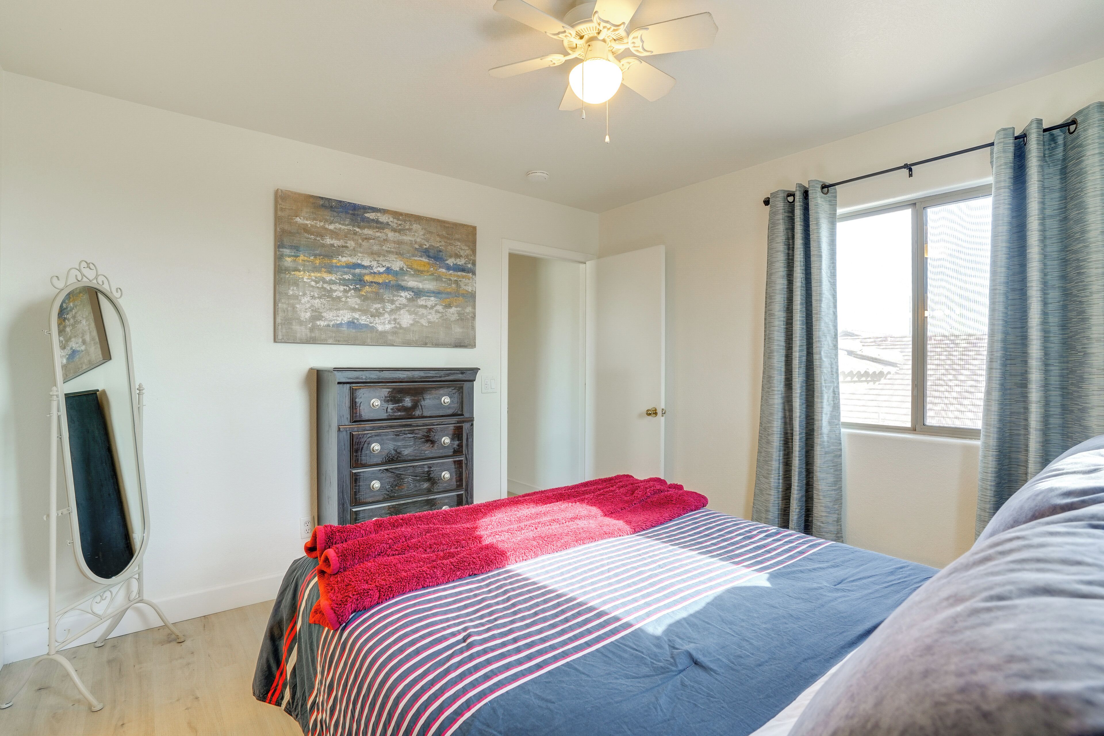 Une chambre à coucher avec un lit, une commode, un ventilateur de plafond, un tableau et une fenêtre avec des rideaux.
