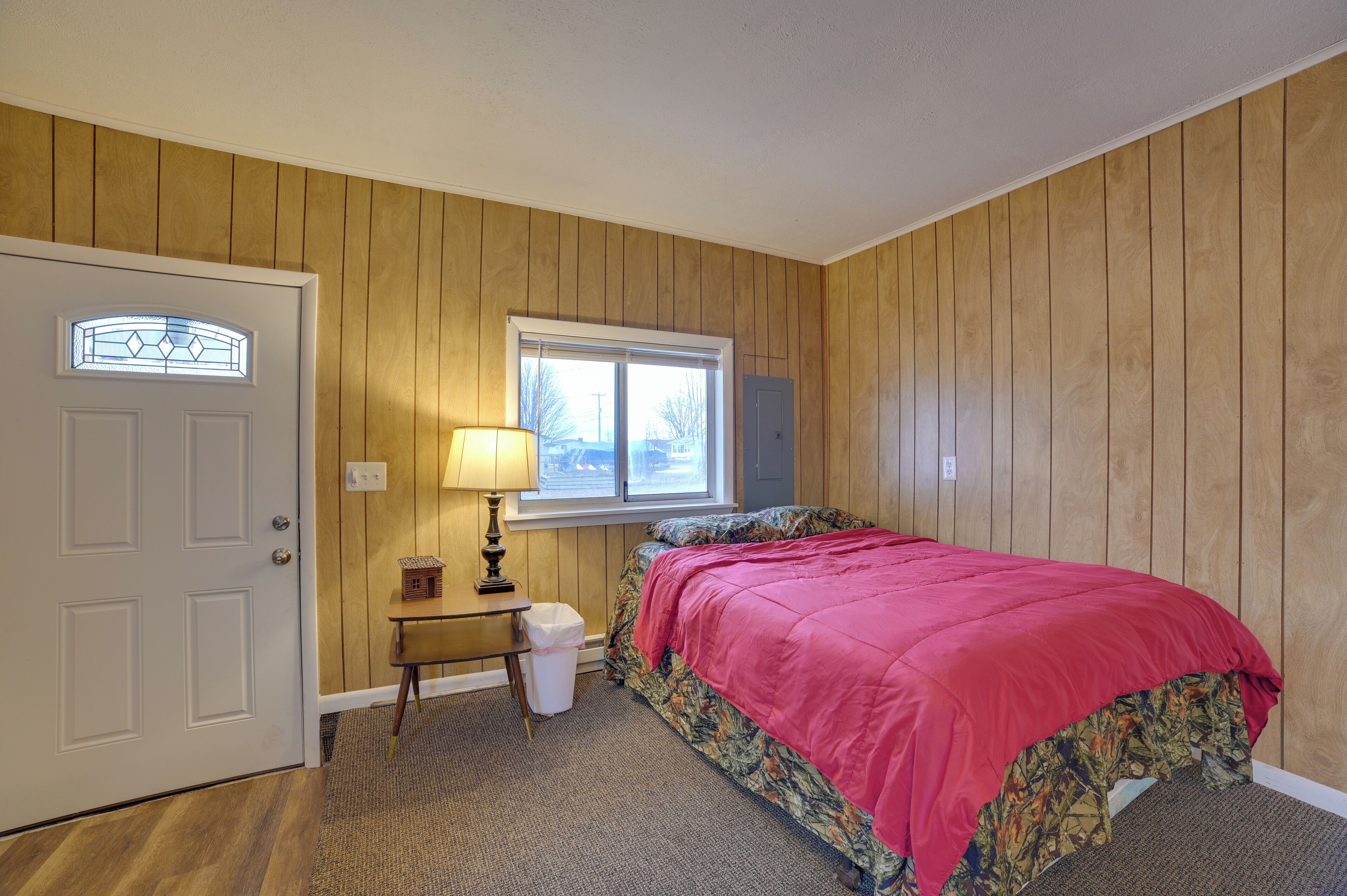 A bedroom with wooden paneling, a bed with a red blanket, a window with blinds, a nightstand with a lamp, and a door.