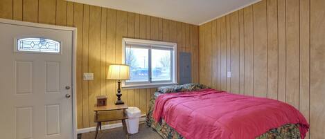 A bedroom with wooden paneling, a bed with a red blanket, a window with blinds, a nightstand with a lamp, and a door.