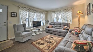 A living room with a grey sofa, a grey armchair, a television on a stand, a fan, and a lamp. - Cozy Houghton Lake Heights Cottage w/ Private Yard (Houghton Lake)