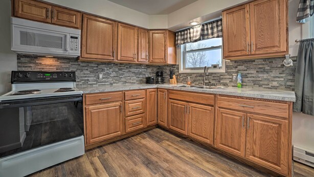 A kitchen with wooden cabinets, a microwave, stove, and a window with curtains. - Cozy Houghton Lake Heights Cottage w/ Private Yard (Houghton Lake)