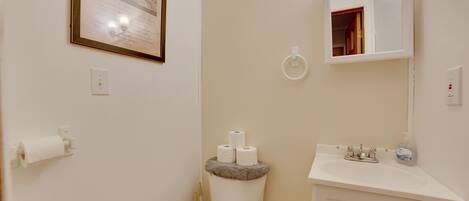 Un baño con inodoro, lavamanos y espejo. Hay un cuadro en la pared y un rollo de papel higiénico en el inodoro.