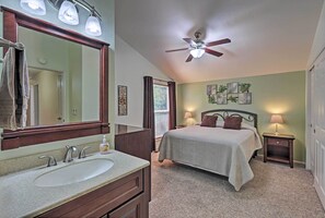 Une chambre à coucher comprenant un lit, des tables de chevet, un ventilateur de plafond, un grand miroir et une salle de bain avec un lavabo et un meuble-lavabo.