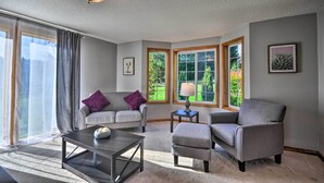 A living room with a sofa, armchair, coffee table, and a lamp. There are large windows with curtains, a ceiling light fixture, and framed pictures on the walls. - Peaceful, Pet-friendly Washougal Paradise! (Washougal)
