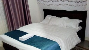 A neatly made bed with white and blue bedding, a dark wooden headboard, and a white curtain. - Apartment Sleeps 6 With Free Parking (Nairobi)