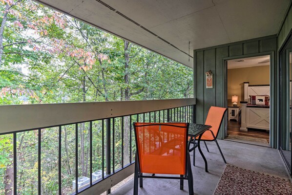 A balcony with a table and chairs, a view of trees, and a kitchen area visible through an open door.