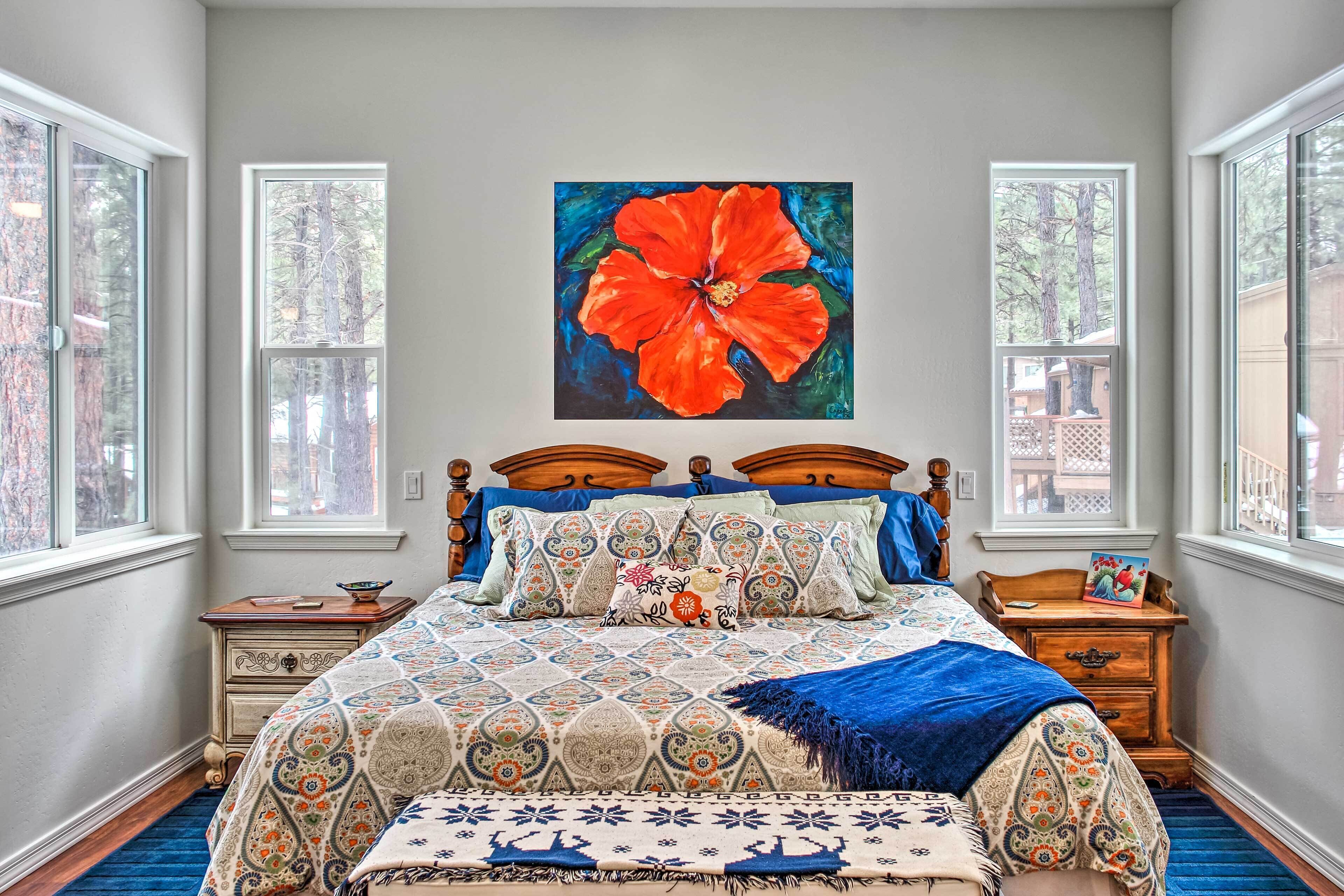 Une chambre à coucher avec un grand lit, deux tables de chevet, une tête de lit en bois, un tableau représentant une fleur rouge aux couleurs vives et de grandes fenêtres.