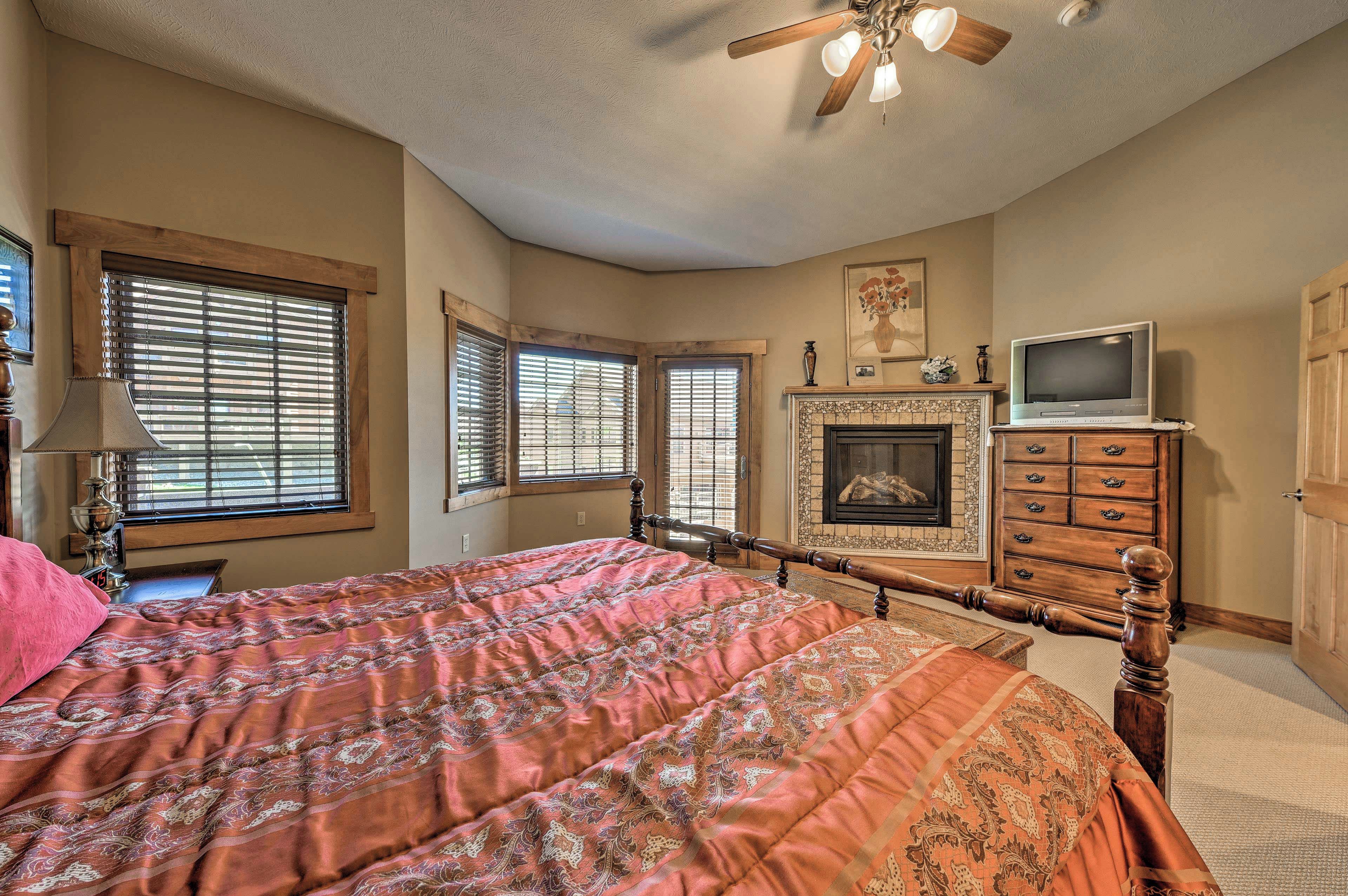 A bedroom with a large bed, a fireplace, a television on a dresser, and a ceiling fan.