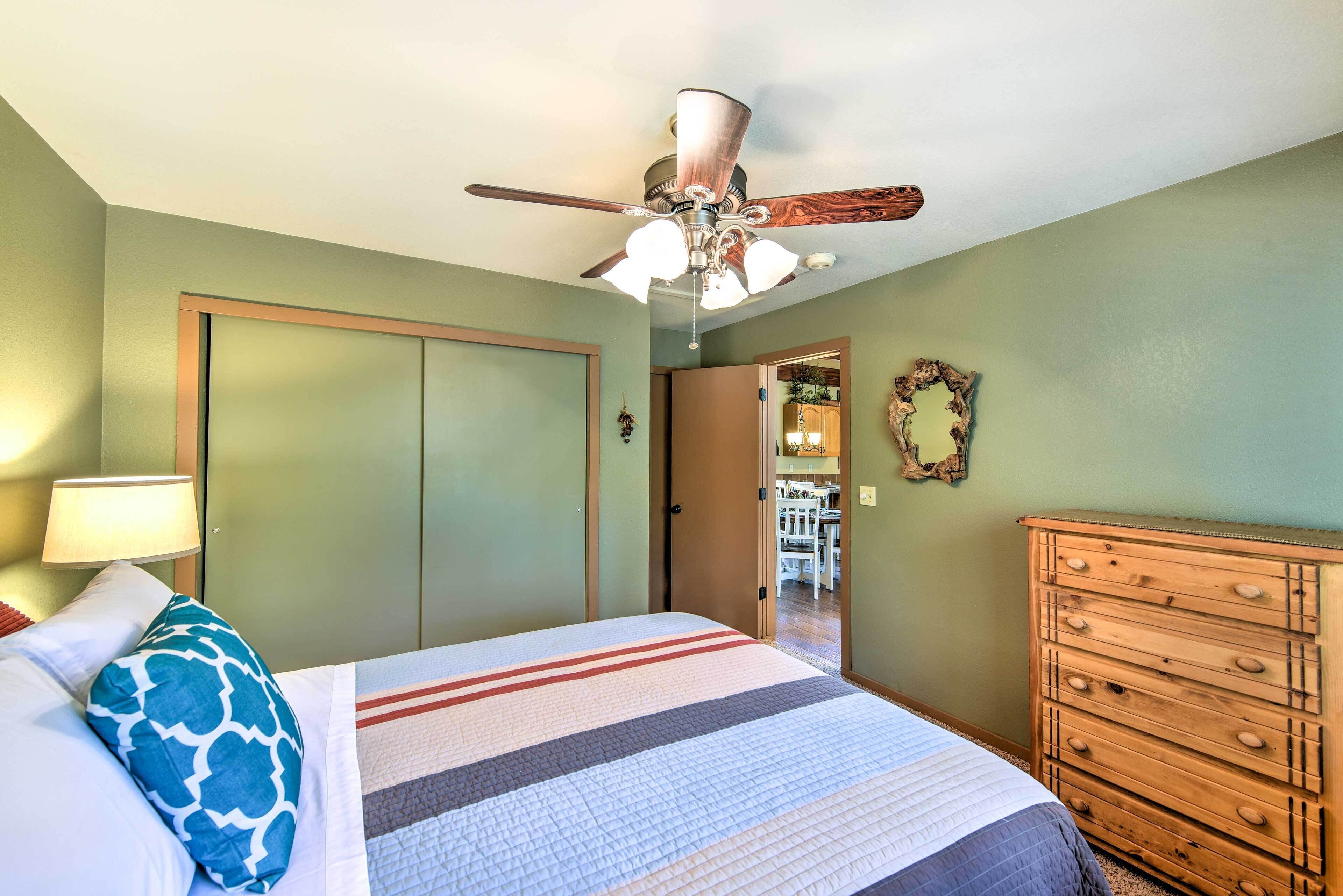 Un dormitorio con cama, cómoda de madera, ventilador de techo y una puerta que da a otra habitación.