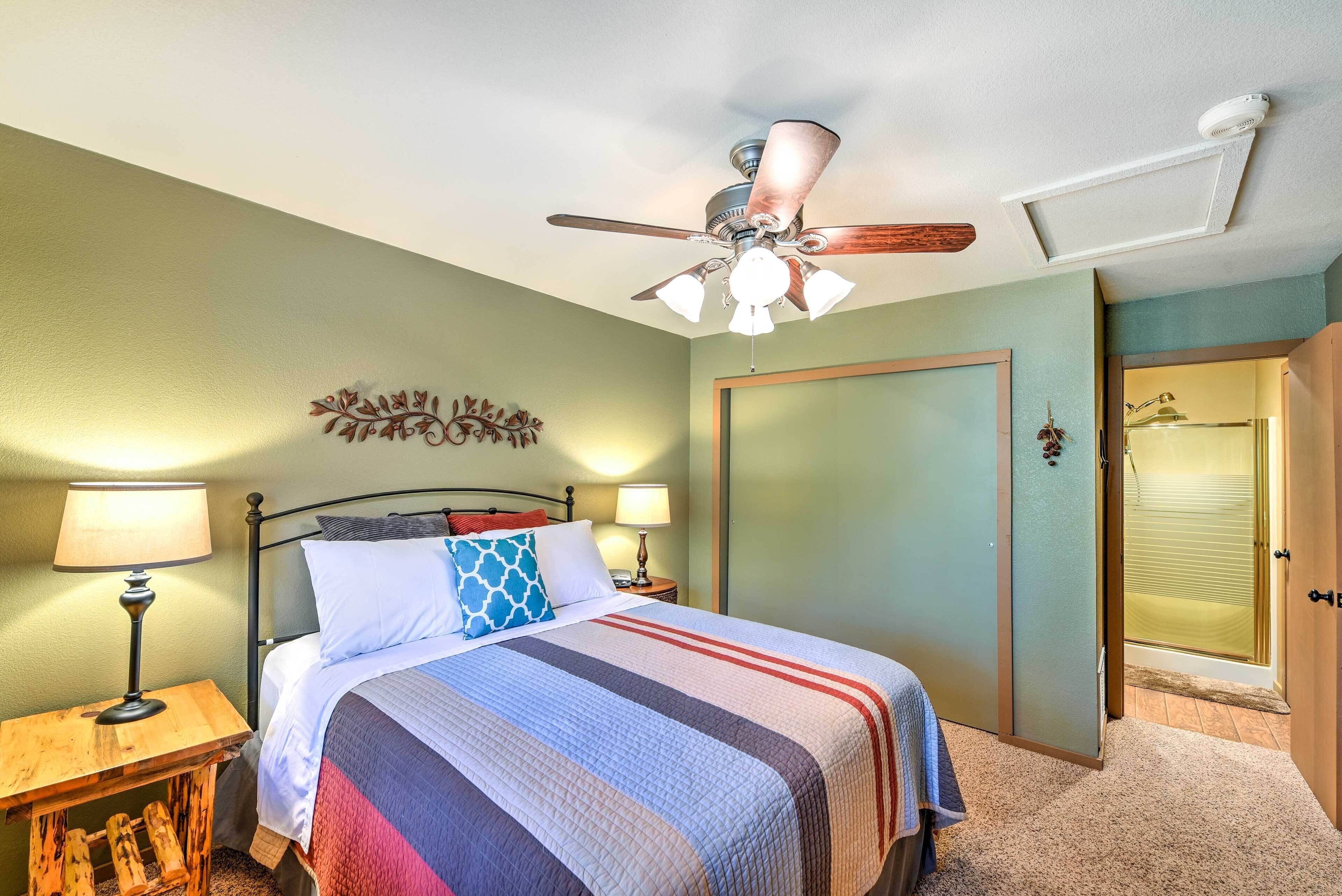 A bedroom with a bed, two bedside lamps, a ceiling fan, a decorative wall piece, and a door leading to a bathroom.