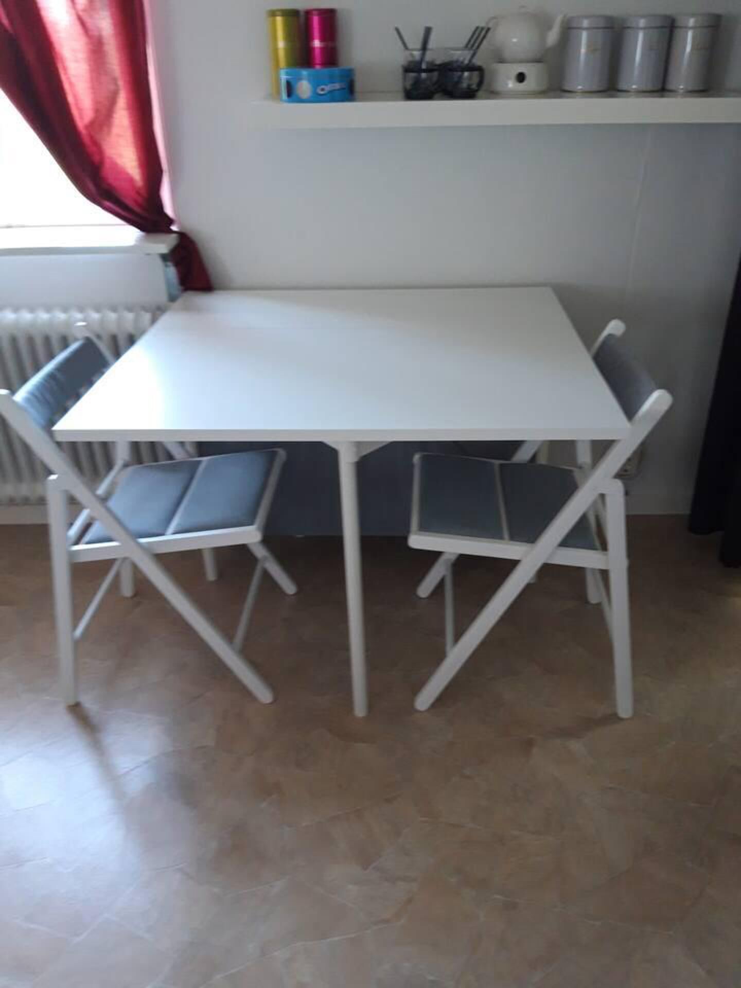 A white table with four chairs, a radiator, and kitchen items on the wall.