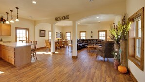 A spacious living area with a central island, a dining area with wooden chairs, a leather sofa, and a 'WELCOME' sign above the archway.