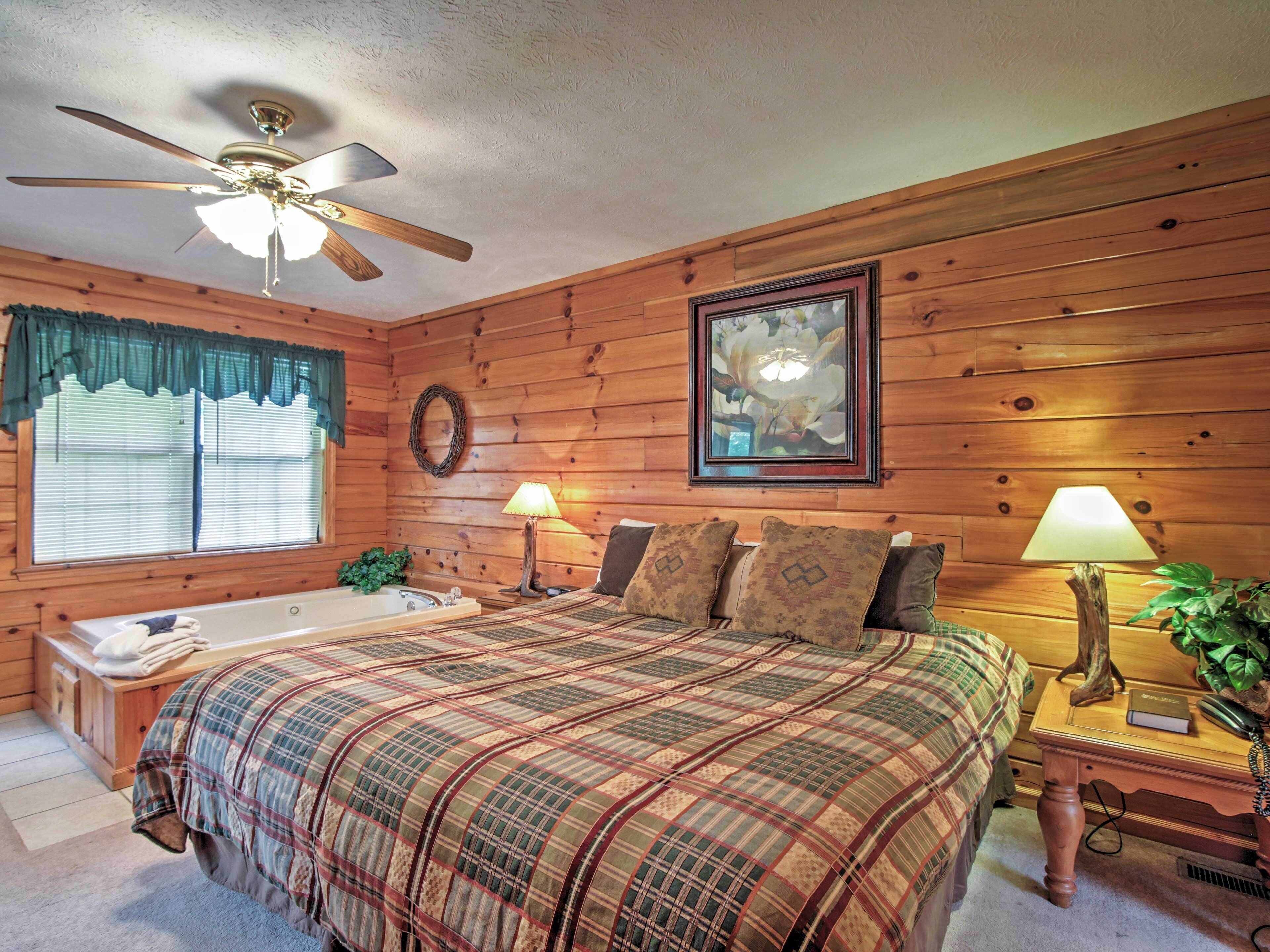 A cozy cabin-style bedroom with a large bed, a ceiling fan, a window with blinds, a framed picture on the wall, and a nightstand with a lamp.