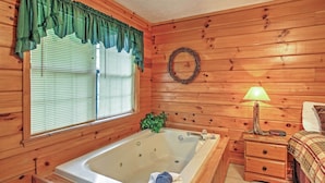 L’intérieur d’un chalet en bois, avec une grande baignoire, une fenêtre avec des stores, une table de chevet avec une lampe et un lit recouvert d’une couverture à carreaux.
