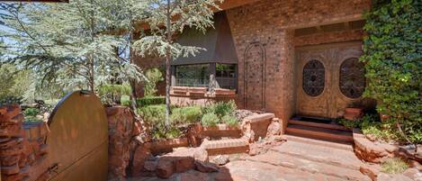 A stone pathway leading to a wooden door with a window, surrounded by greenery and a brick wall.