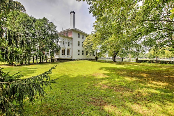 Um edifício grande e branco com uma chaminé alta, cercado por árvores verdes exuberantes e um gramado bem cuidado.