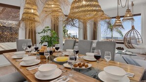 A dining area with a long wooden table set for a meal, surrounded by grey chairs. There are hanging wicker chandeliers, a potted plant, and a view of the beach through large windows. - KARAT Villa Uchova - Cho Pancho (El Frontón)