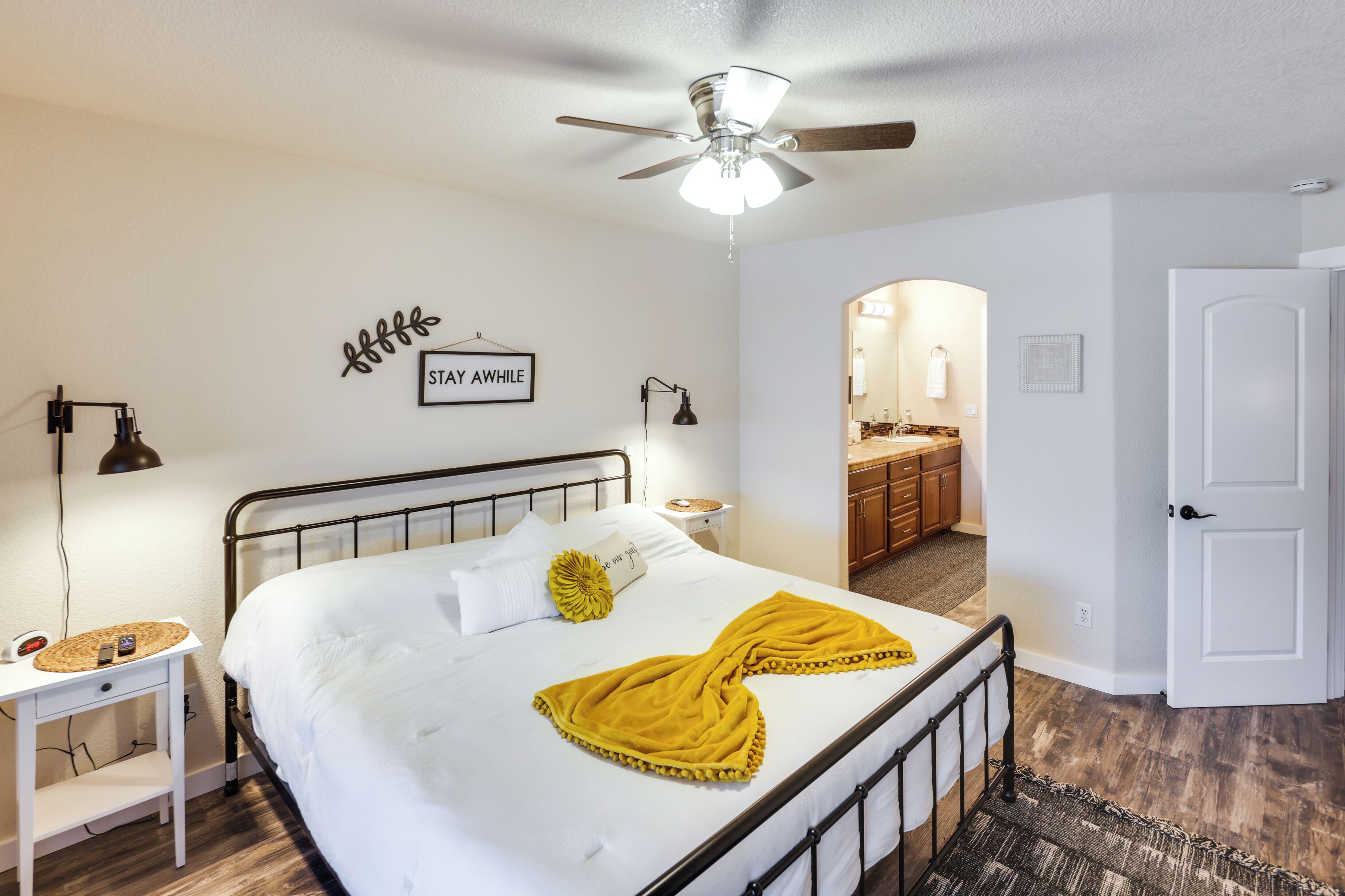 Un dormitorio con una cama, ventilador de techo, mesita de noche y mesilla.