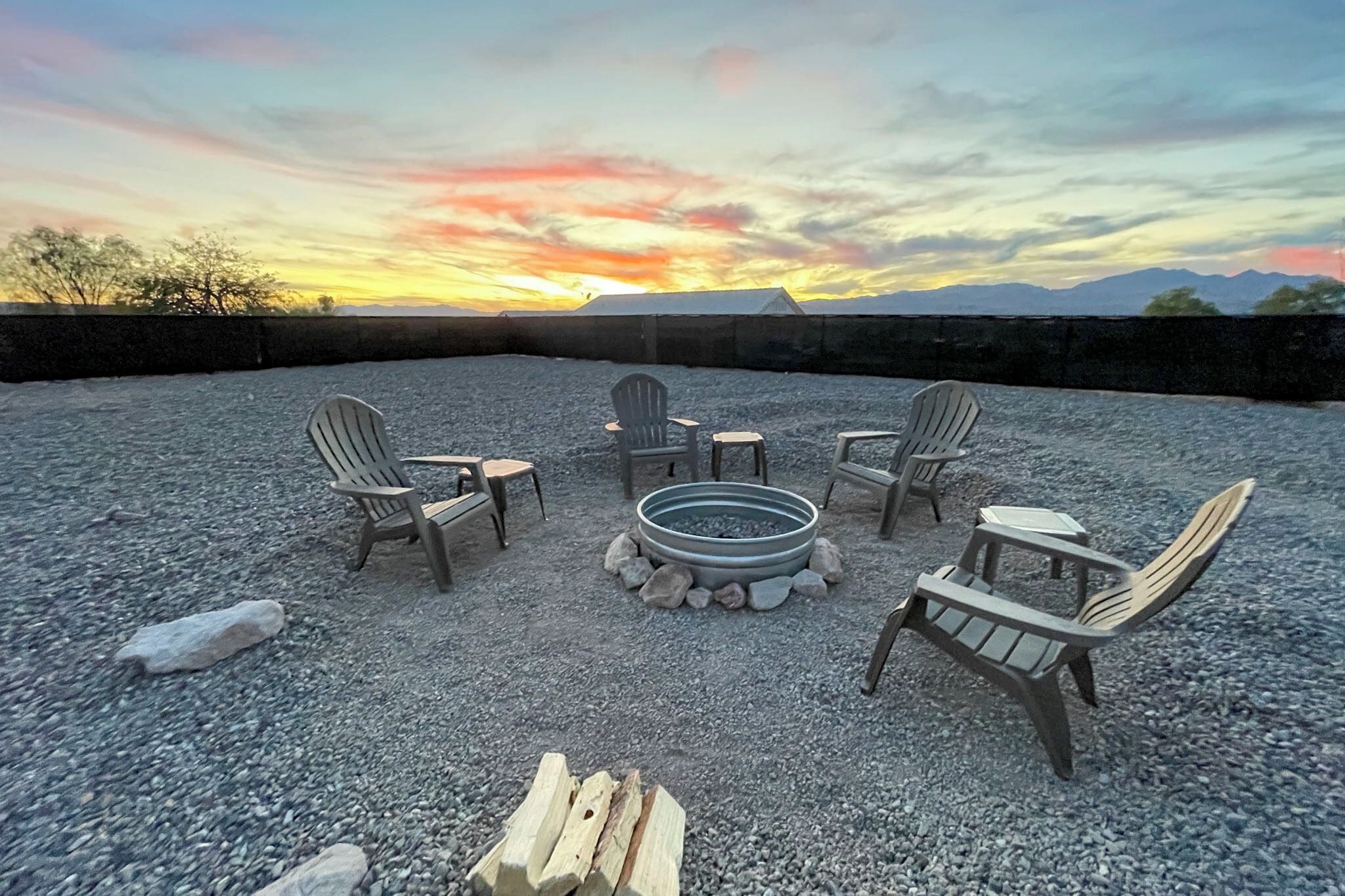 Un patio en la azotea con cuatro sillas Adirondack dispuestas alrededor de un hogar circular, sobre un suelo de grava y con una vista panorámica del atardecer de fondo.