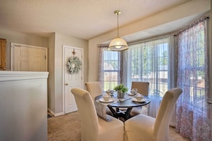 A dining area with a round table set for four, surrounded by cushioned chairs, a hanging light fixture, and a view through large windows with sheer curtains.