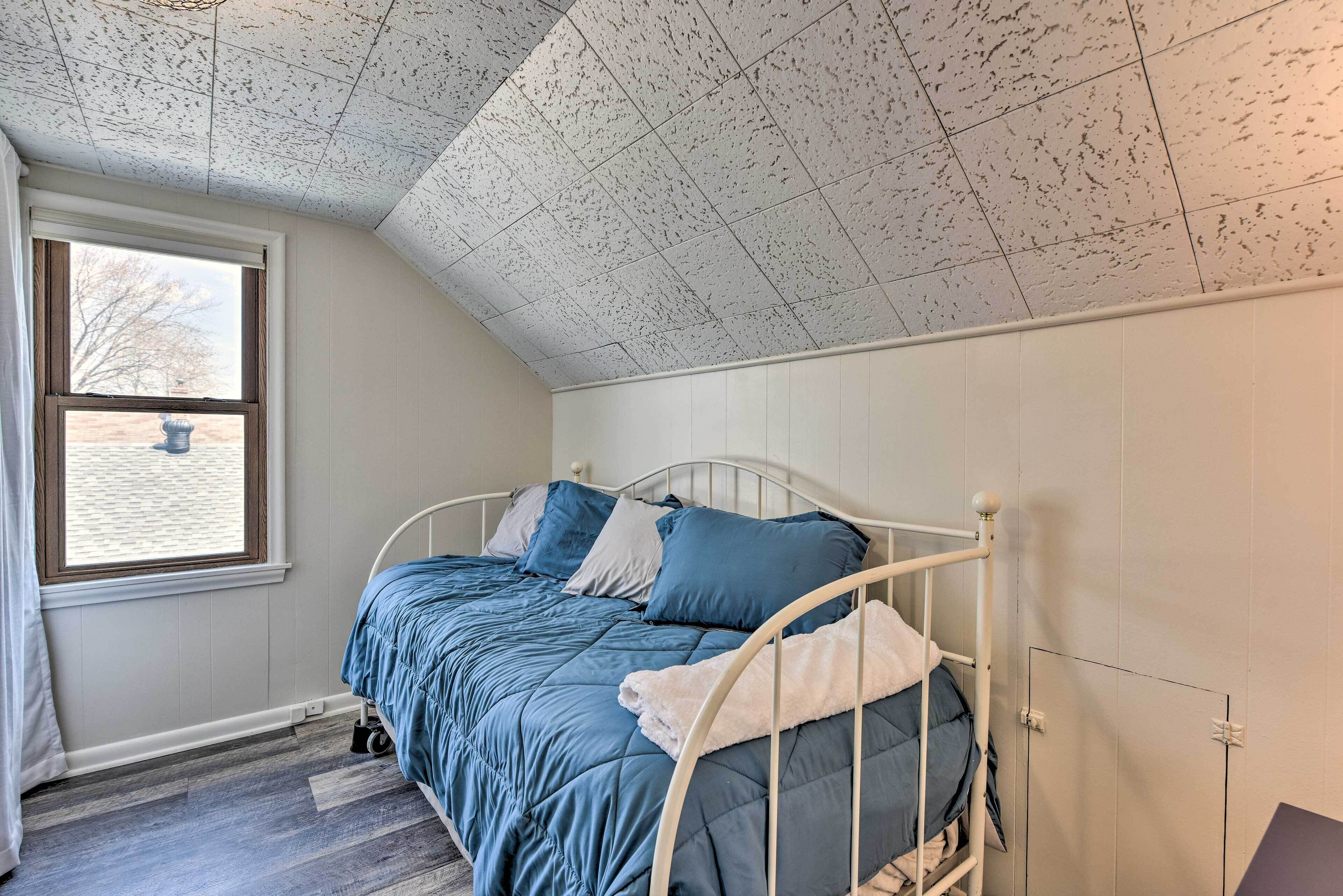A bedroom with a sloped ceiling, a bed with blue bedding, a window with curtains, and a white door.