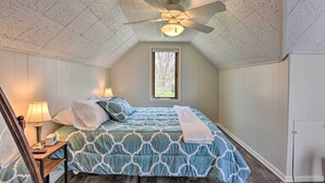 Un dormitorio con techo inclinado, ventilador de techo, cama con colcha estampada, mesita de noche con lámpara y ventana con vista a árboles.