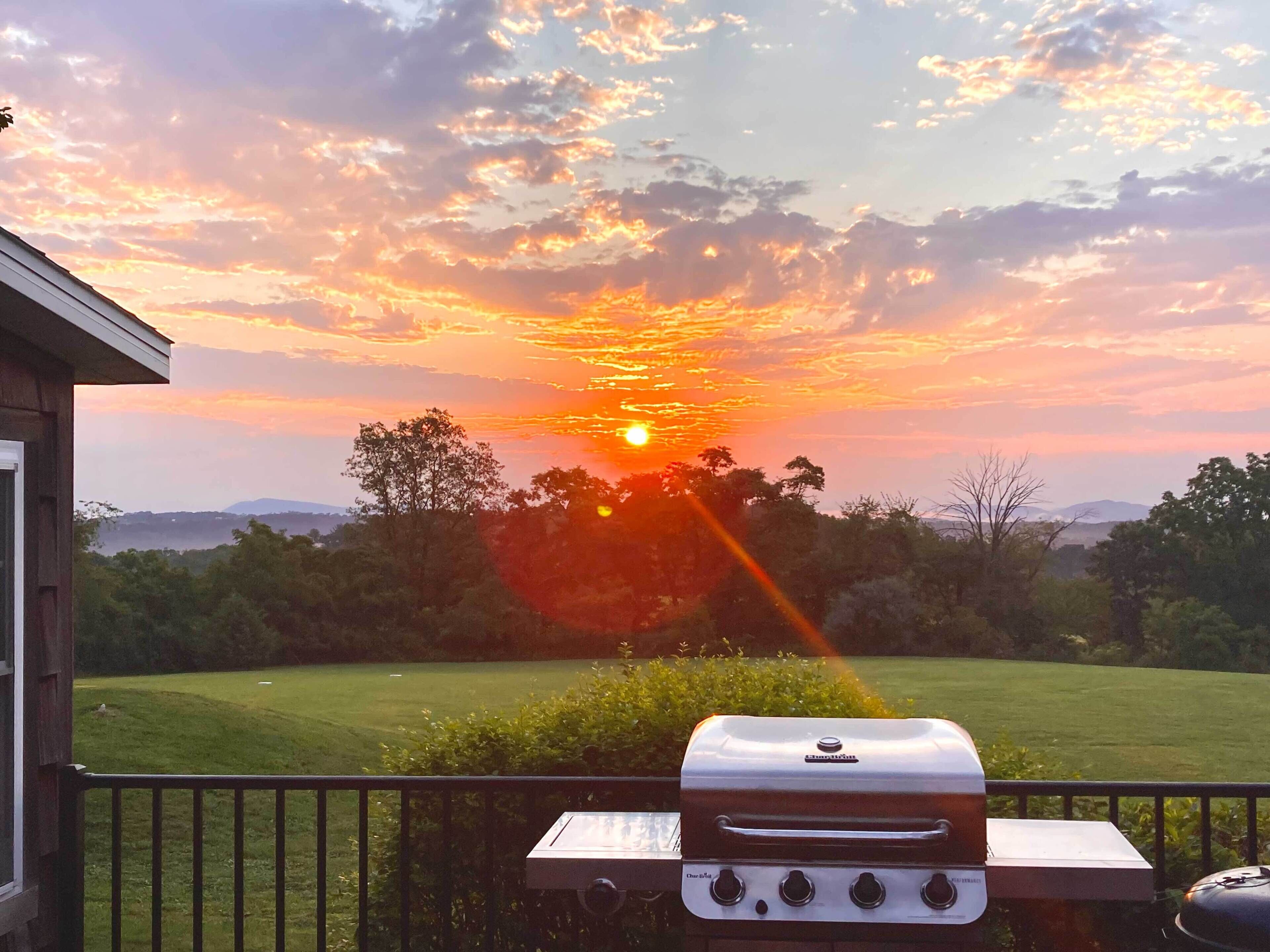 A barbecue grill on a balcony overlooking a grassy landscape with trees and a sunset in the background.