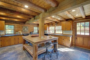 A log cabin kitchen with a large island, wooden cabinets, and stone flooring. - Cozy Log Cabin Getaway w/ Fire Pit + 3 Acres! (Ruckersville)