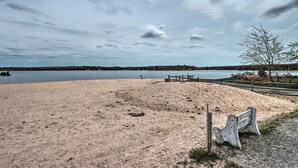 Une plage de sable avec un banc en bois et une clôture, donnant sur une étendue d’eau calme et sur des arbres situés au loin.