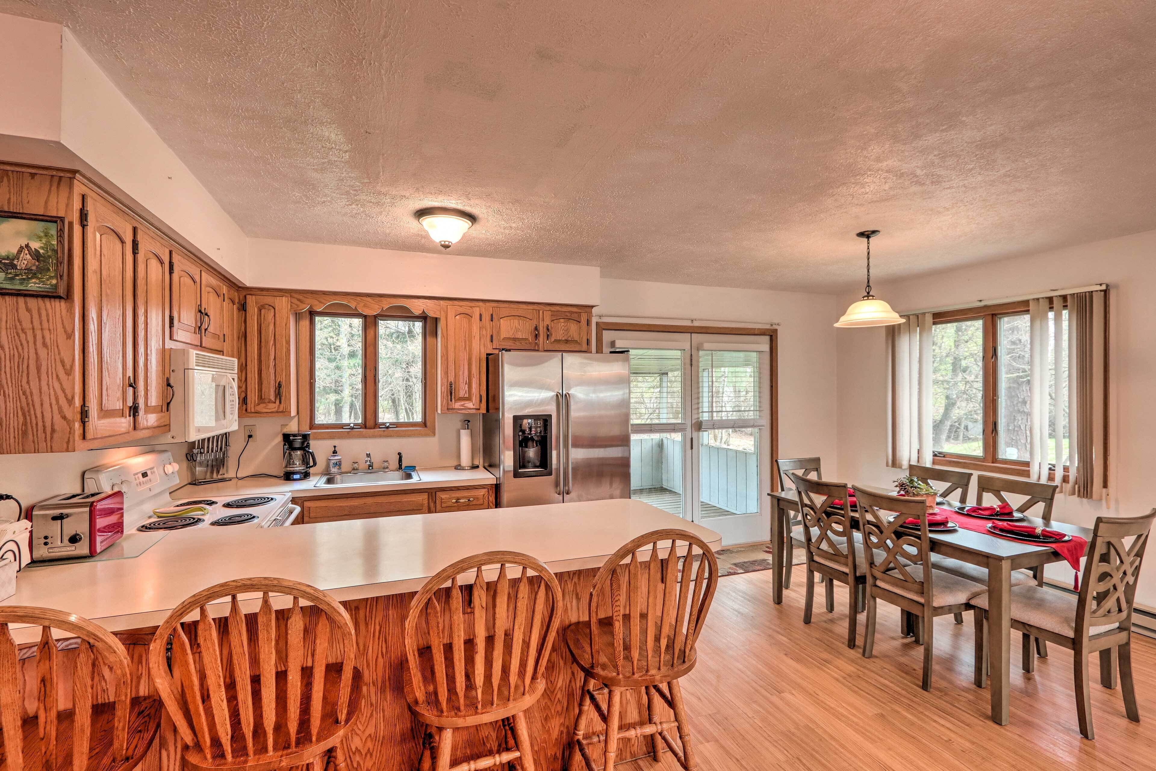 A spacious kitchen with wooden cabinets, a large island with bar stools, a dining table with chairs, and a view of the outdoors through large windows.
