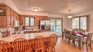 Une cuisine spacieuse avec des meubles en bois, un grand îlot central doté de tabourets, une table à manger avec des chaises, et une vue sur l’extérieur grâce à de grandes fenêtres.