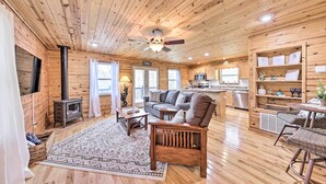 A cozy cabin interior with a living room, kitchen, and dining area, featuring wooden floors and walls, a fireplace, and a ceiling fan. - Scenic Murphy Escape With Hot Tub & Fire Pit! (Murphy)