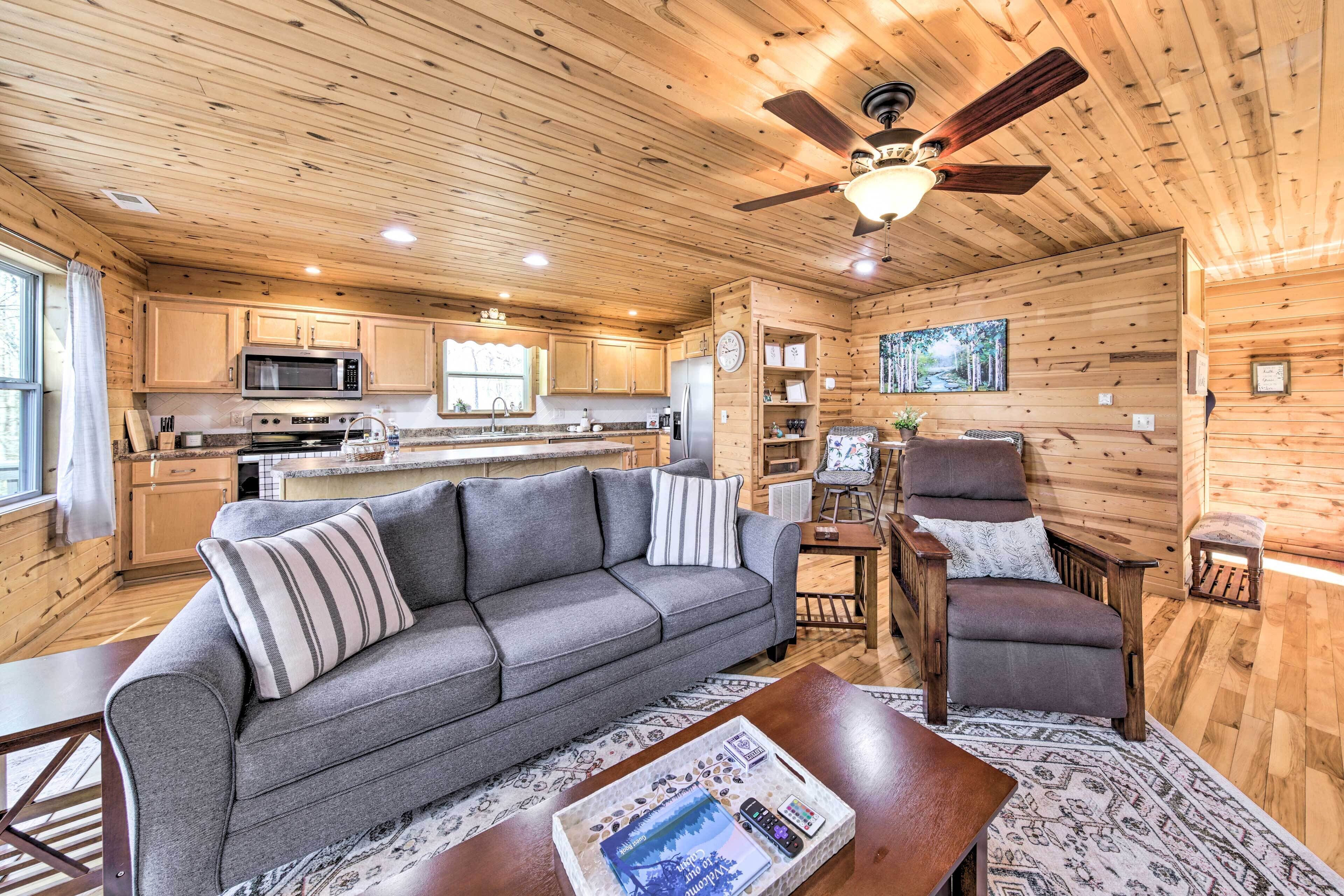 A cozy cabin interior with a wooden ceiling, a sofa, a chair, a dining table, and a kitchen area.