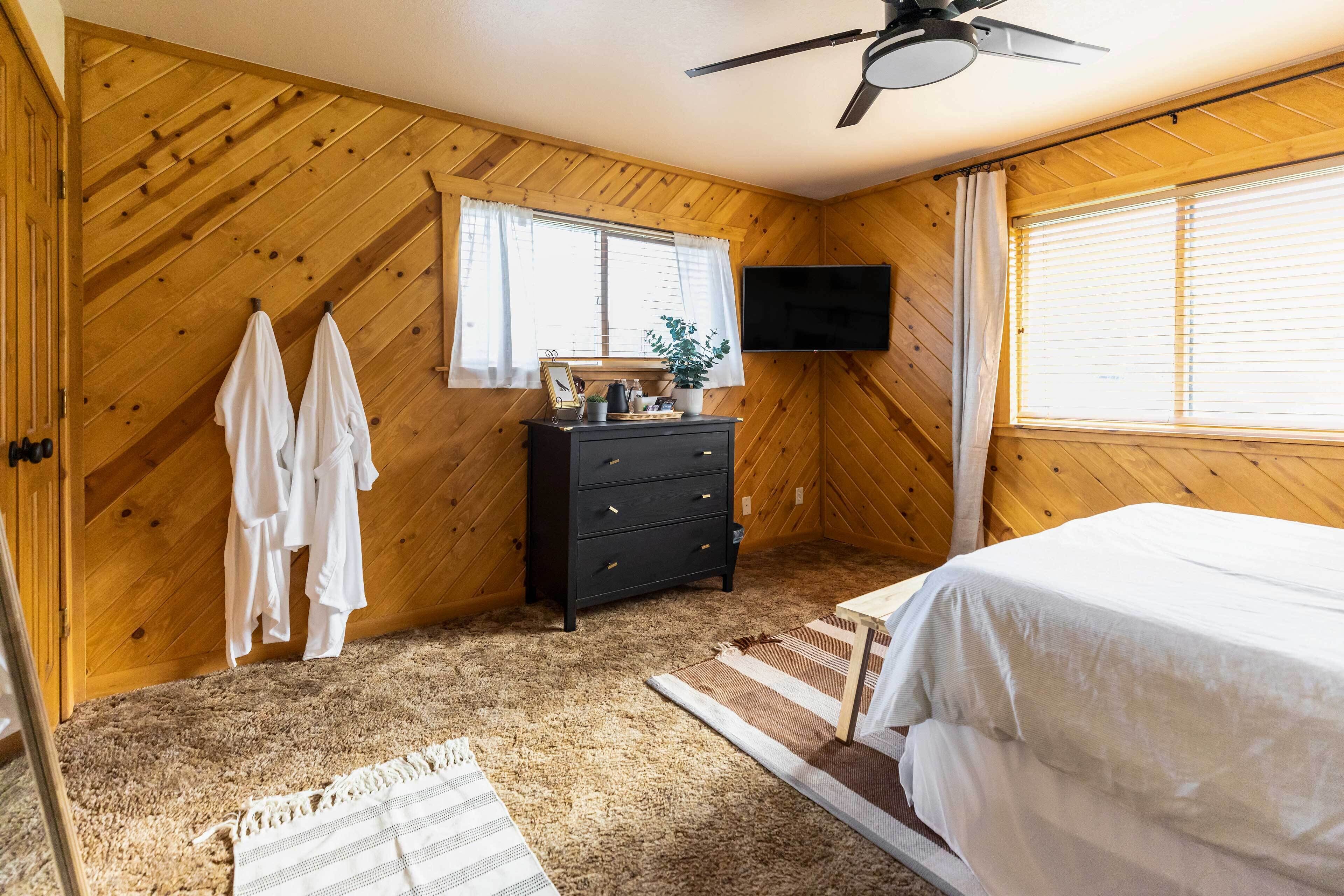 Une chambre avec des murs en bois, un lit, une commode, un téléviseur et une fenêtre avec des rideaux.